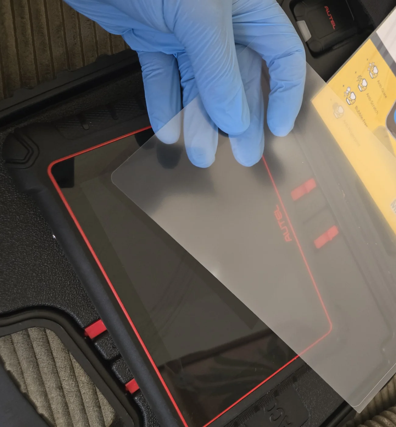 Przezroczysta matowa folia ochronna trzymana w niebieskiej rękawiczce nitrylowej nad ekranem tabletu diagnostycznego Autel leżącego w otwartej walizce transportowej, przymiarka przed naklejeniem