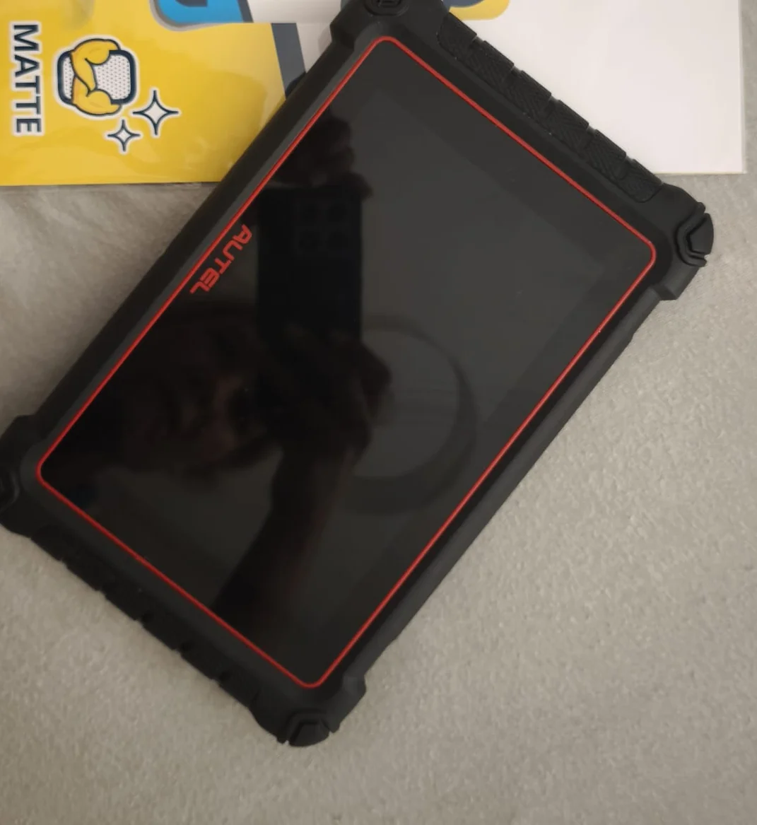 Tablet diagnostyczny Autel w czarnej wzmocnionej obudowie z czerwoną ramką na jasnym blacie po naklejeniu matowej folii ochronnej, ekran wygaszony bez pęcherzy powietrza