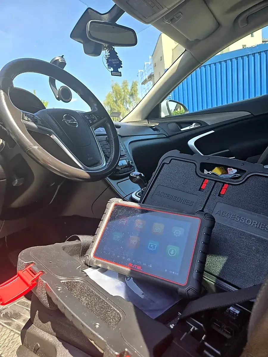 Tester Autel MaxiCOM MK900-BT na fotelu Opla Insignia obok otwartej walizki z akcesoriami, ekran z menu diagnostycznym