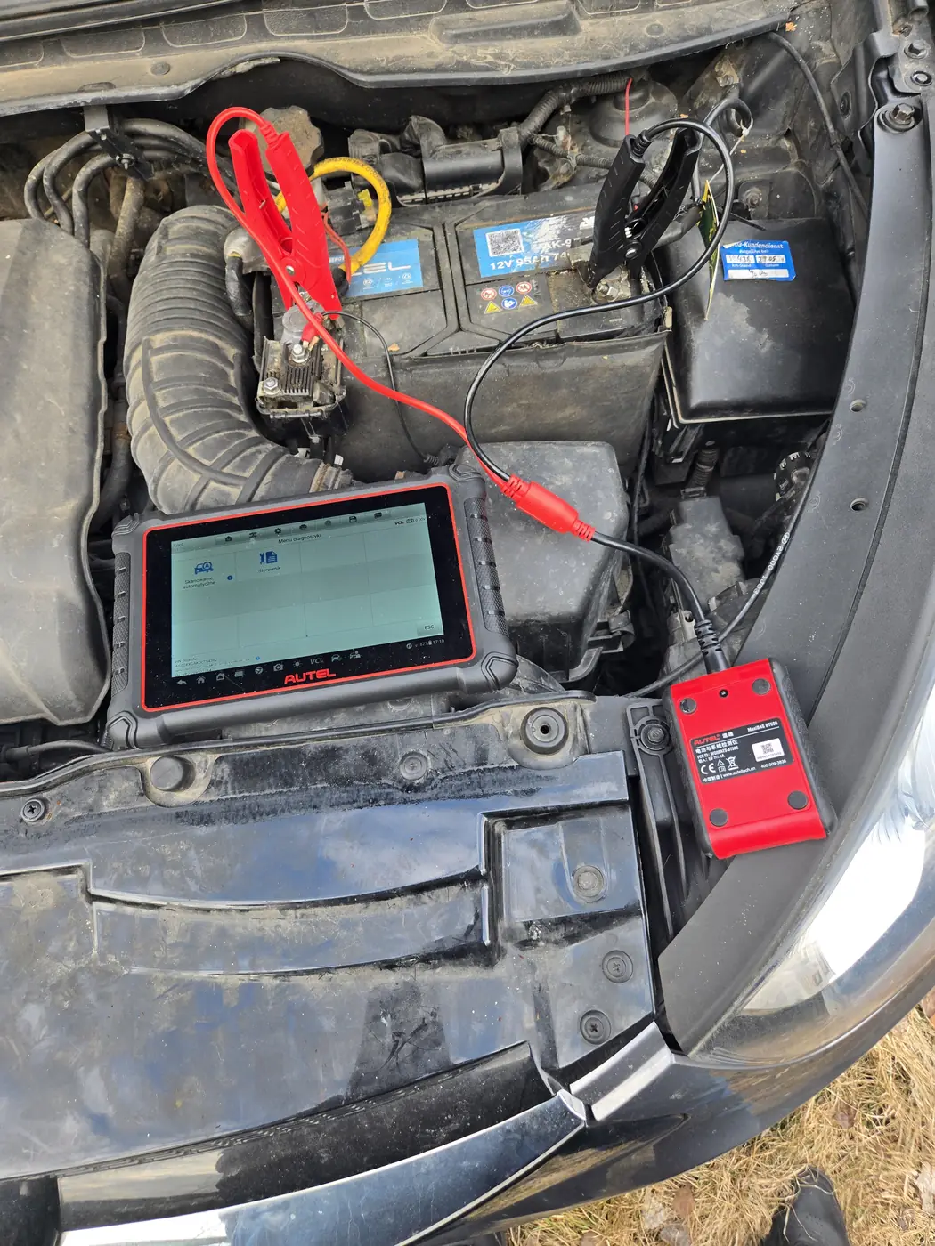 Test akumulatora i alternatora testerem Autel BT506 pod maską auta — moduł z klipsami na biegunach i tablet diagnostyczny w komorze silnika